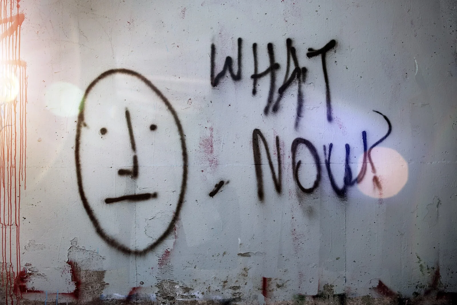 Graffiti on a concrete wall: a blank drawn face beside the words WHAT NOW?